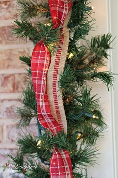 a christmas tree with red and white plaid ribbon hanging from it's branches, next to a brick wall