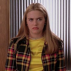 a woman in a yellow shirt and plaid jacket standing next to a wooden door with vertical blinds