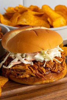 a pulled pork sandwich with coleslaw and cheese on a cutting board next to potato chips