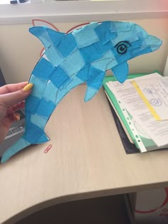 a hand is holding a piece of paper that looks like a blue fish on a desk