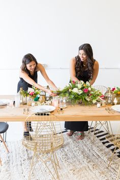 two women sitting at a wooden table with flowers and greenery on it, preparing to cut the cake