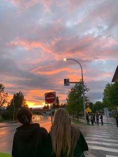 two people are sitting on a bench watching the sun go down at an intersection with traffic lights and street signs