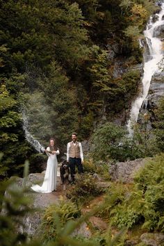 a man and woman standing in front of a waterfall with a dog on their lap