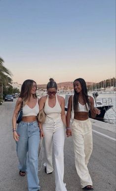 three women are walking down the road near some water and palm trees, one has her hand in her other's pocket