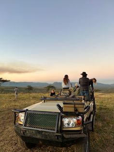 people are sitting in the back of a truck on top of a grassy field with mountains in the background