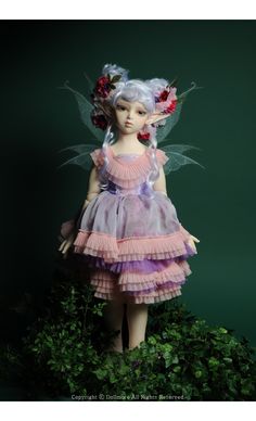 a doll dressed in pink and purple is standing on top of some green bushes with flowers