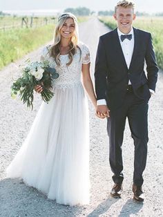 a bride and groom walking down a dirt road holding hands, dressed in tuxedos