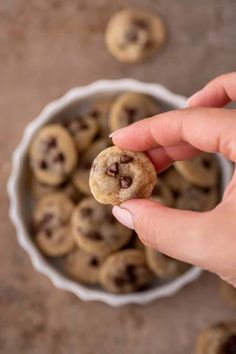 a hand holding a chocolate chip cookie in front of a bowl full of mini cookies