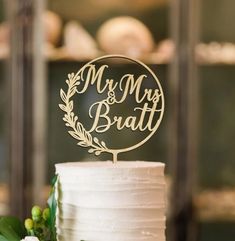 a white cake topped with a wooden mr and mrs topper next to greenery