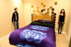 a man and woman standing in front of a bed with purple bedspread on it