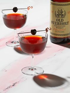 two glasses filled with red liquid next to a bottle of alcohol on a white table