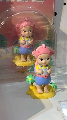 two figurines are on display in a glass case, one is wearing a pink hat and the other is holding a flower