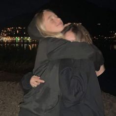 two women hugging each other in front of a body of water at night with buildings in the background