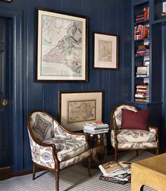 two chairs and a table in a room with books on the shelves behind them are framed maps