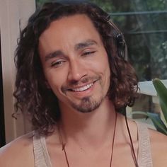 a man with long hair wearing headphones smiles at the camera while standing in front of a window