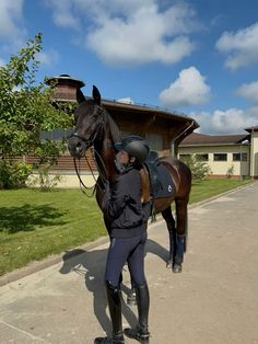 a woman in riding gear standing next to a brown horse on a sidewalk near a building