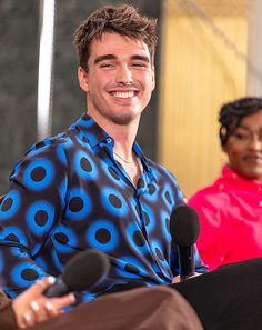 a smiling man sitting in front of microphones next to another person wearing a blue and black shirt