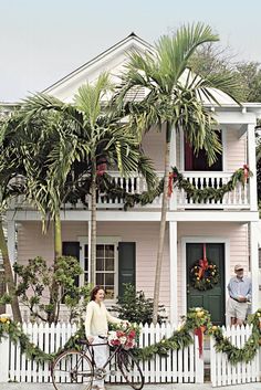 two people standing in front of a pink house with wreaths and palm trees on the fence