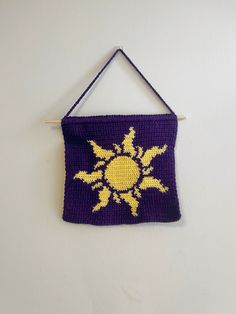 a crocheted wall hanging with a sun motif in purple and yellow on a white background