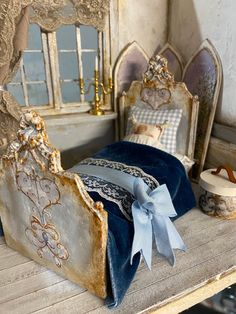 a dollhouse bed with blue sheets and pillows on it's headboard, sitting next to a window