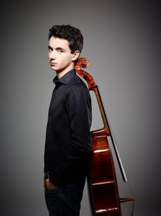 a young man holding a cello in his right hand and standing against a gray background