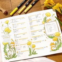 an open planner sitting on top of a wooden table next to yellow flowers and markers