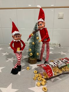 two elfs are standing next to a christmas tree and bag of cheerios on the floor