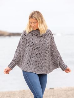 a woman is walking on the beach wearing a gray knitted ponchy sweater