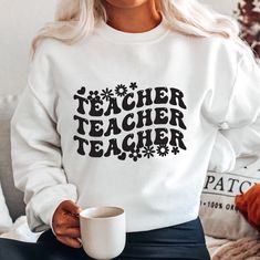 a woman sitting on a couch holding a coffee cup and wearing a sweatshirt that says teacher teacher