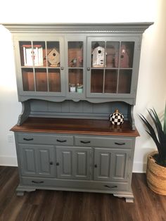 a gray china cabinet with glass doors and drawers on the top, next to a potted plant