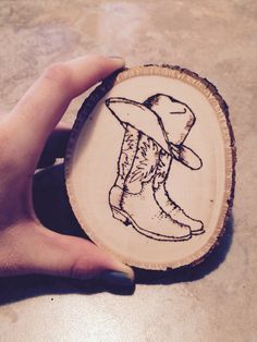 a hand is holding a small wood slice with a drawing of a cowboy boot on it