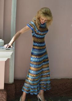 a woman in a blue and brown dress leaning against a pink wall with her hand on the ground