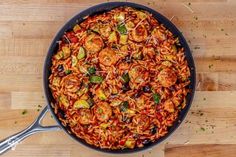 a skillet filled with pasta and vegetables on top of a wooden table next to utensils