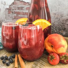 two glasses filled with red liquid next to fruit and cinnamon sticks on a table near a pitcher