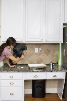 a woman in a kitchen with white cabinets and black trash can on the counter top