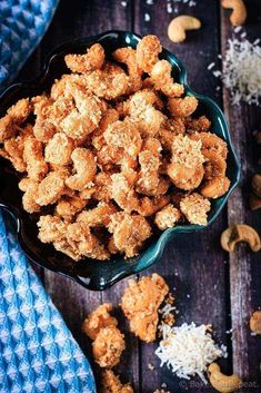 a bowl filled with fried chicken next to cashews on a blue towel and wooden table