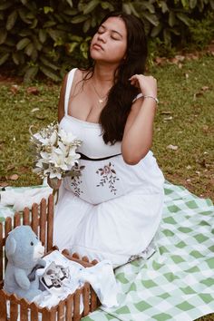 a woman in white dress sitting on blanket next to teddy bear and basket with flowers