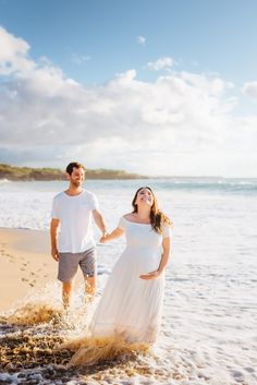 a pregnant couple walking along the beach in front of the ocean holding hands and smiling