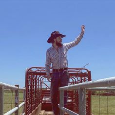 a man standing on top of a metal fence next to a cow pen with his hand in the air