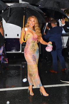 a beautiful woman holding an umbrella while standing on a wet street with other people in the background