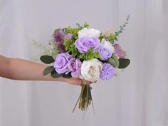 a person holding a bouquet of flowers in their hand with white, purple and green foliage