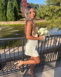 a beautiful woman in white dress holding flowers by a railing with trees and water behind her