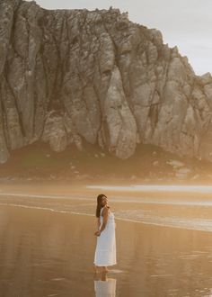 a woman standing on the beach in front of a rock formation with her reflection in the wet sand