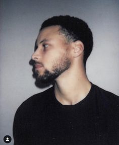 a man with a beard and black shirt looking off to the side in front of a gray wall