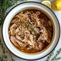 a white bowl filled with meat and garnish next to lemons, parsley and herbs