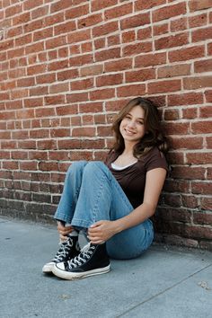 a woman sitting against a brick wall with her feet on the ground and looking at the camera