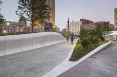 two people walking down a paved walkway in the middle of a city with tall buildings