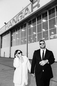 a man and woman walking in front of a hanger with their cell phones to their ears