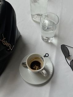 a cup of coffee on a saucer next to sunglasses and a black bag with the word louis written on it