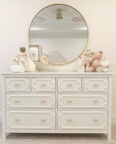 a white dresser topped with lots of stuffed animals and a round mirror above it's top